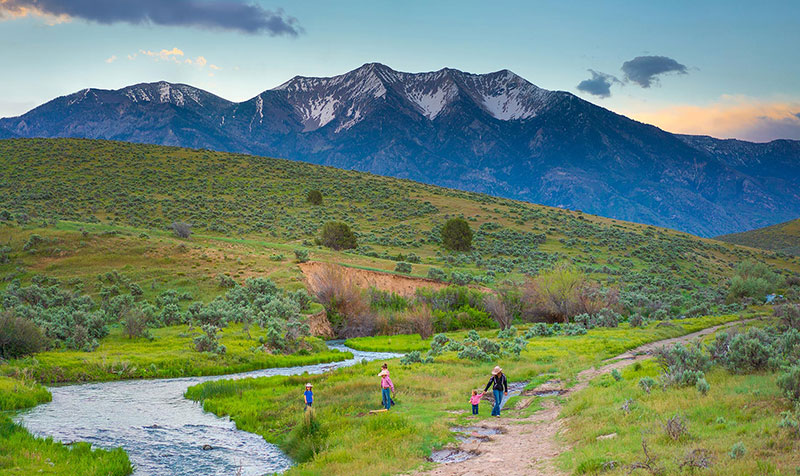 Family walking near a river with a mountain backdrop.